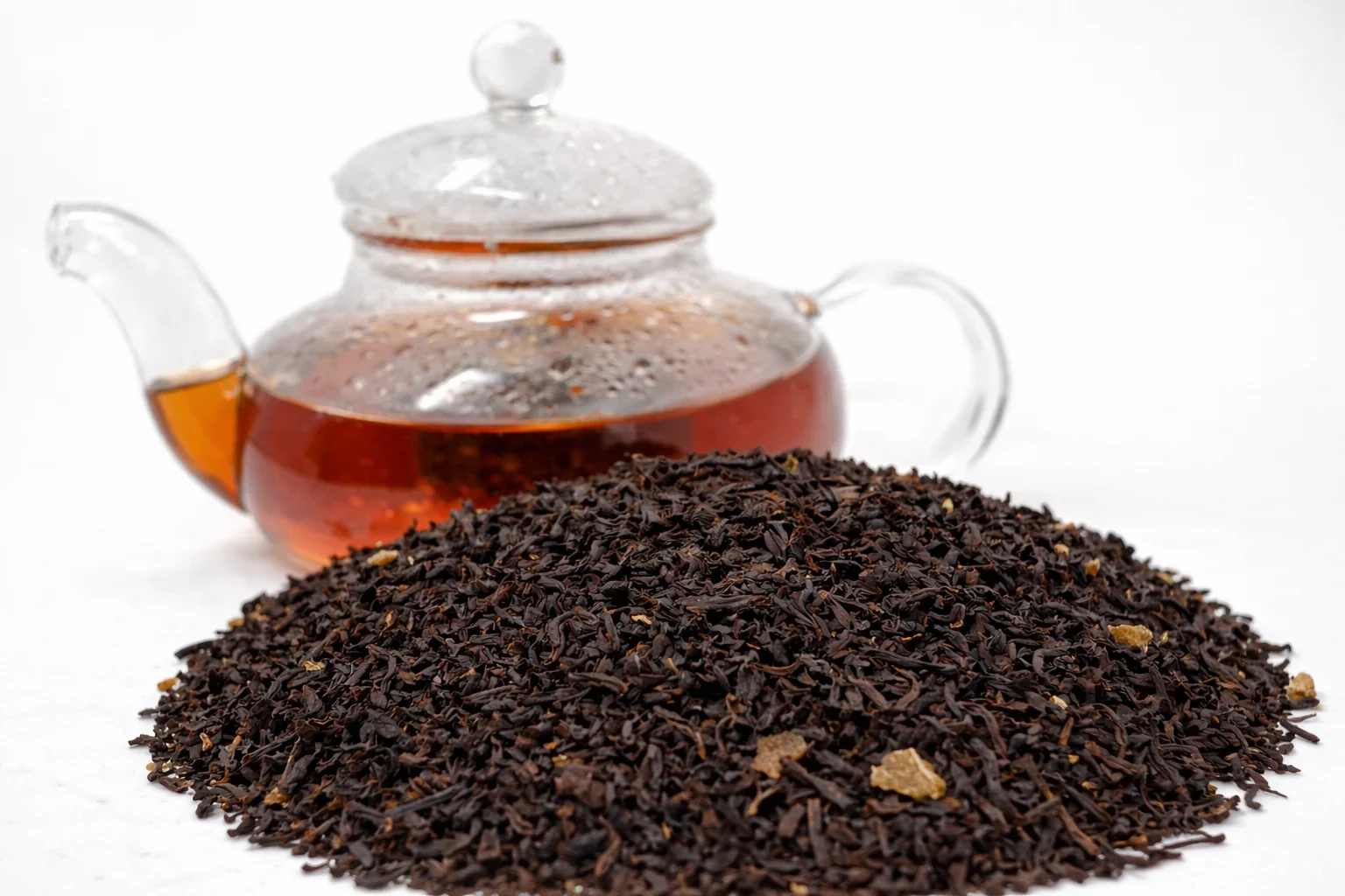 Glass teapot with tea and a pile of black tea leaves on a white background